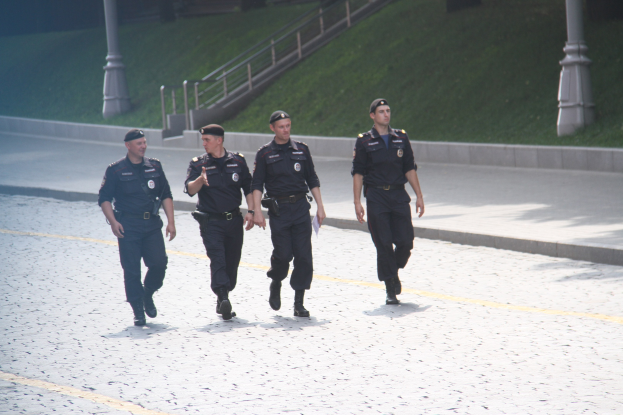 Drei Polizisten in Uniform und Mützen, die eine Straße mit Masten, Geländern, Treppen und Gras im Hintergrund entlanggehen.