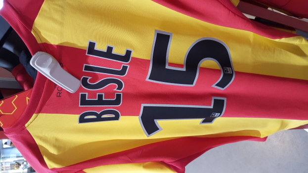 Ein roter und gelber Fußballtrikot mit Text und Zahlen hängt an einem Ständer in einem Geschäft, mit anderen Gegenständen und dem Hintergrund im Hintergrund.