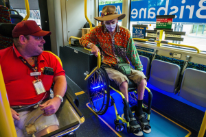 Zwei Männer im Rollstuhl in einem Bus, einer trägt ein rotes T-Shirt und eine rote Mütze, der andere ein orangefarbenes Hemd und einen braunen Hut, beide tragen Masken und Ausweise, mit gelben Stangen und Glasfenstern, die Bäume, Gebäude und den Himmel zeigen.