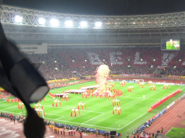 Ein Stadion mit Flutlicht, Eisengrills, Zuschauern, stehenden Personen, Werbetafeln, Anzeigetafeln, Sicherheitskamera, Fahnen und Dekorationselementen.