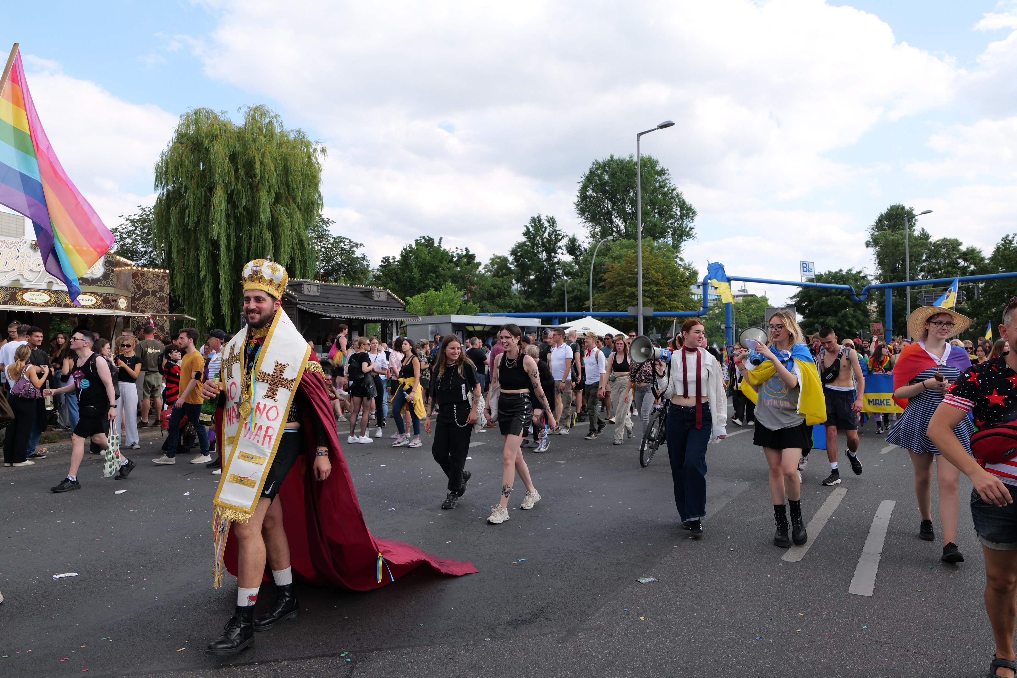 Eine Gruppe von Menschen marschiert bei der Pride Parade 2018 mit einer Regenbogenflagge und Musikinstrumenten, während im Hintergrund Laternenpfähle, Bäume und Hütten zu sehen sind.