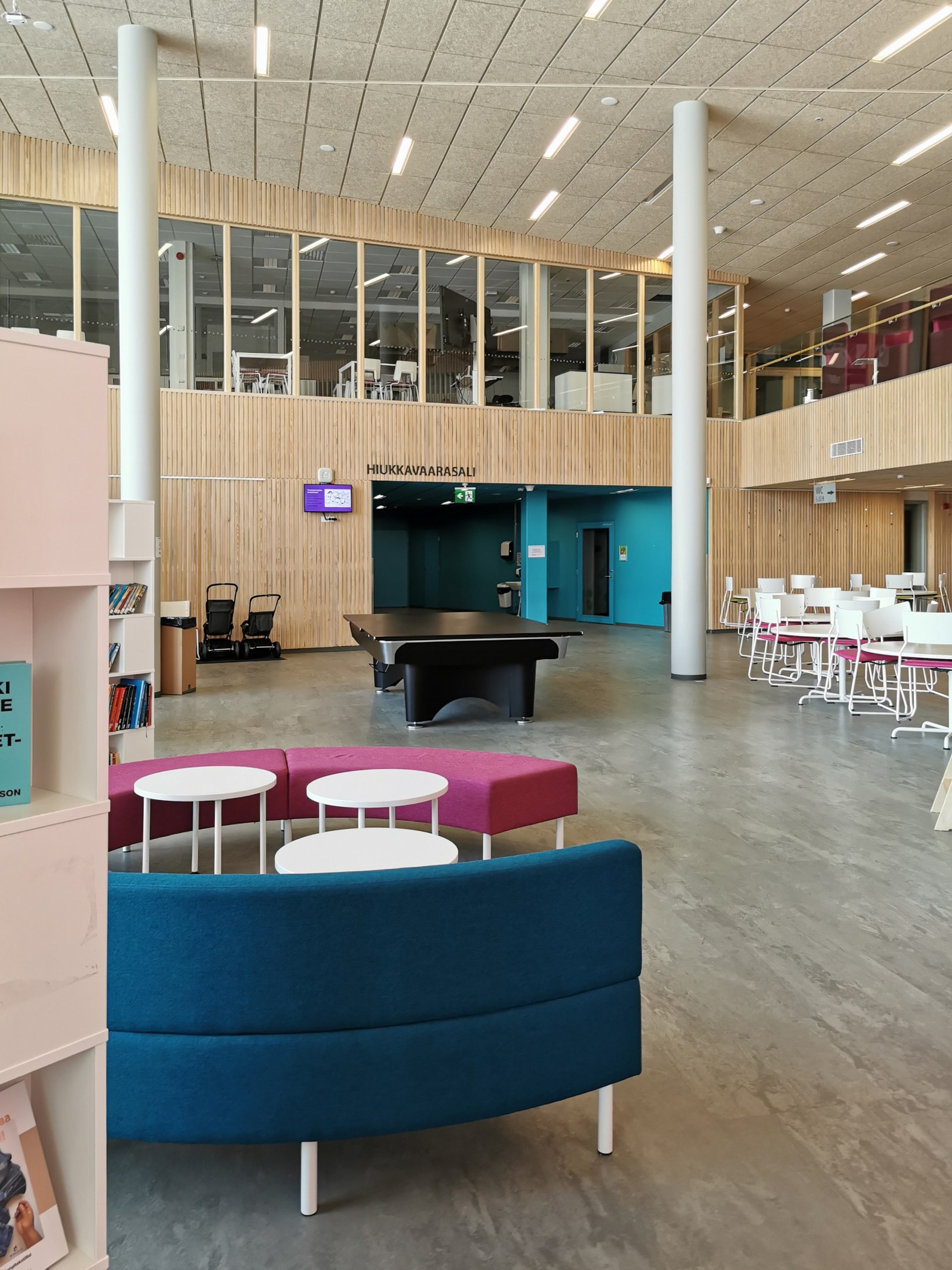 Ein geräumiger Raum ähnlich einer Schulbibliothek mit Tischen, Stühlen, Sofas, Regalen, Säulen, Deckenleuchten und einem Wandfernseher.