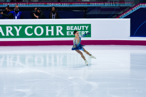 Eine Frau in einem blauen Kleid fährt auf einem Eisstadion Schlittschuh, während die Menge Kameras hält und ein Schild mit der Aufschrift 'Tessa Virtue und Scott Moir' im Hintergrund zu sehen ist.