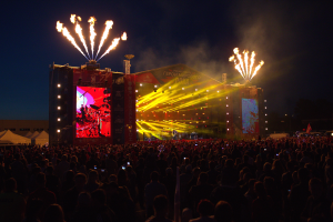 Eine große Menschenmenge auf einem Musikfestival mit einer Bühne in der Mitte, Musiker, die unter hellen Scheinwerfern spielen, Banner mit Text, Zelte links und ein dunkler Himmel mit Feuerwerk im Hintergrund.