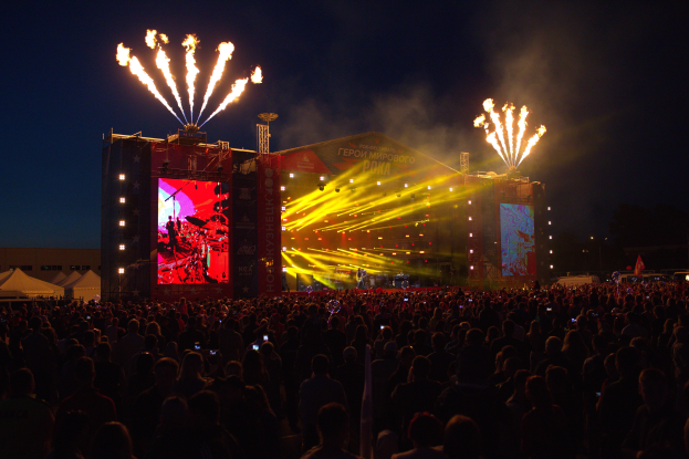 Eine große Menschenmenge auf einem Musikfestival mit einer Bühne in der Mitte, Musiker, die unter hellen Scheinwerfern spielen, Banner mit Text, Zelte links und ein dunkler Himmel mit Feuerwerk im Hintergrund.