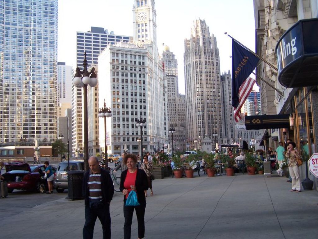 Eine belebte Straßenszene mit Fußgängern, Blumentöpfen, Autos, Straßenlaternen, Gebäuden, Geschäften und Fahnen auf der rechten Seite.