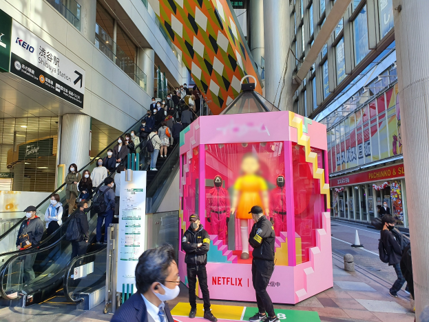 Eine Gruppe von Menschen steht vor einem Gebäude mit Glaswänden und Texttafeln, einige tragen Masken, während eine Rolltreppe mit Menschen zu sehen ist; ein pinker Kasten in der Mitte zeigt Schaufensterpuppen und trägt den Text "Netflix's new Netflix-Boot in Tokyo."