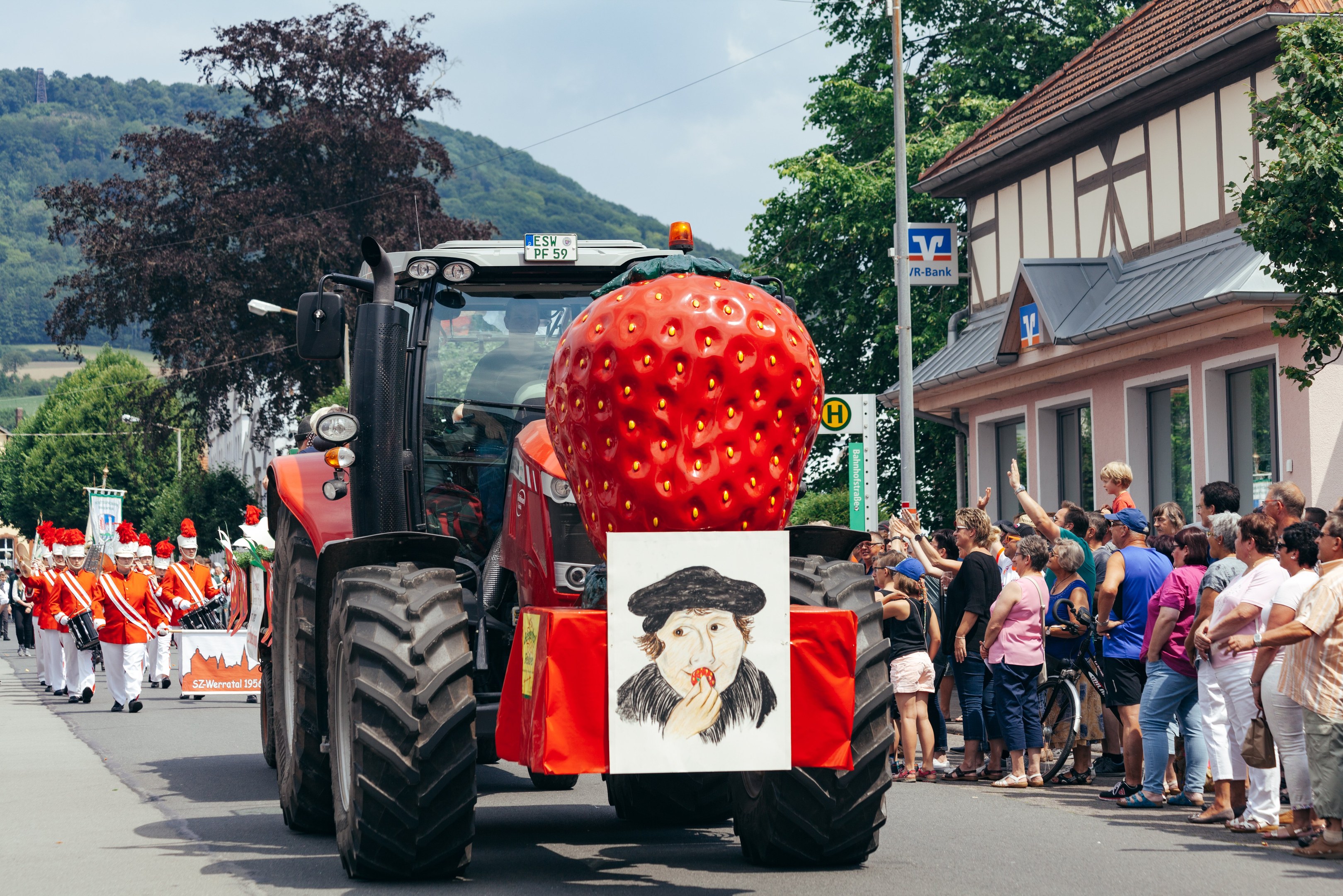 Ein großer Erdbeer-förmiger Schwimmer auf einem Traktor fährt eine Straße entlang an einer Gruppe von Menschen vorbei, die Fahrräder auf dem Gehweg halten, mit Gebäuden, Bäumen und Hügeln im Hintergrund unter einem klaren blauen Himmel.
