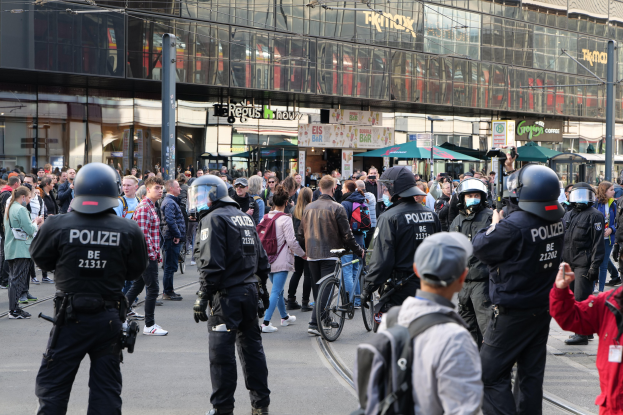 Eine Gruppe von Polizisten vor einer Menge auf der Straße, einige mit Helmen und Taschen oder Fahrrädern, sowie Gebäude mit Glaswänden und Beschriftungen im Hintergrund.