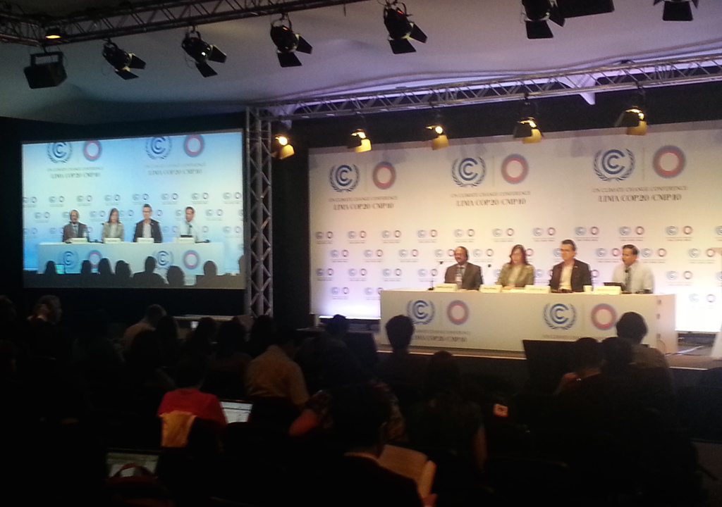 Menschen sitzen auf Stühlen auf einer Bühne mit Namensschildern und Mikrofonen auf einem Tisch davor, während Zuschauer in Stühlen sitzen und die Bühne anschauen, mit einem Projektionsschirm und Beleuchtung auf Ständern im Hintergrund.