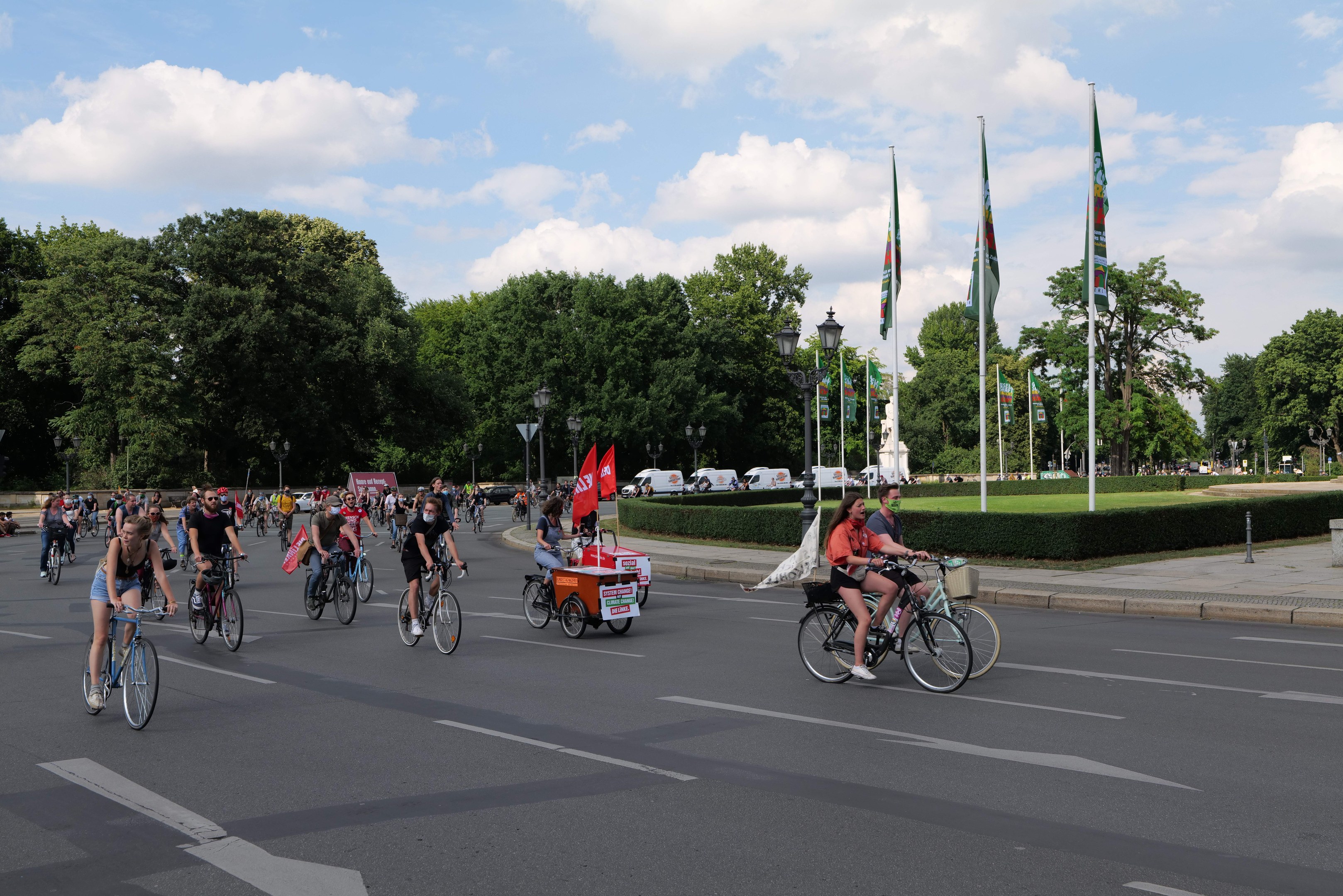 Eine Gruppe von Menschen, die auf Fahrrädern eine von Bäumen gesäumte Straße mit Laternenmasten und Flaggenmasten entlangfahren, während der Pariser Fahrradtag 2016 stattfindet, mit bewölktem Himmel und einigen Fahrzeugen im Hintergrund.