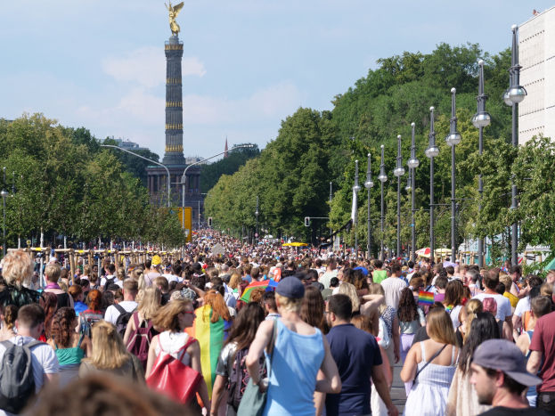 Eine große Menschenmenge mit Taschen geht eine Straße in Berlin, Deutschland, entlang, gesäumt von Laternenmasten, Bäumen, einem Turm mit einer Statue oben drauf, Gebäuden und einem bewölkten Himmel im Hintergrund.
