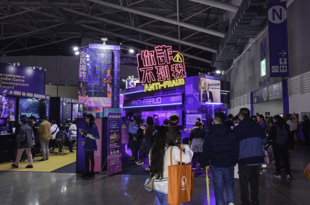 Eine große Gruppe von Menschen steht vor einem Messestand, der als "Indie Game Development Award 2019" gekennzeichnet ist, wobei einige Taschen und verschiedene Anzeigen, Bildschirme und Lichter im Hintergrund zu sehen sind.