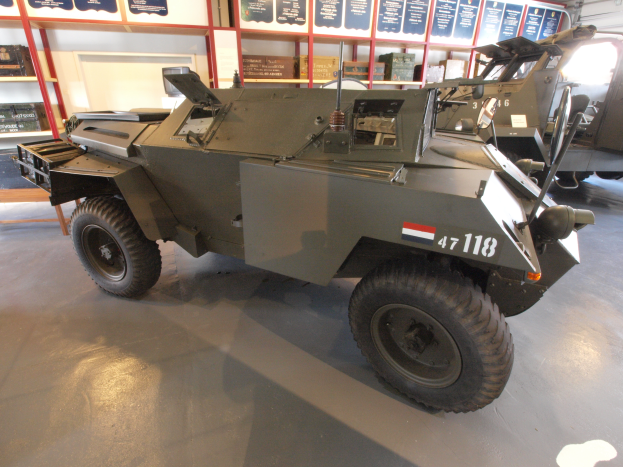 Ein gepanzerter Personentransporter steht in einem Gebäude, umgeben von anderen Fahrzeugen, mit Texttafeln, Regalen, einer Tür und Deckenlampen im Hintergrund.