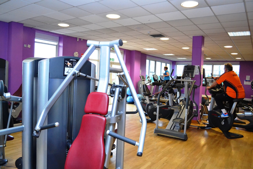 Ein Fitnessstudio mit verschiedenen Geräten und einigen Menschen beim Training, mit einer Decke und Lampen oben