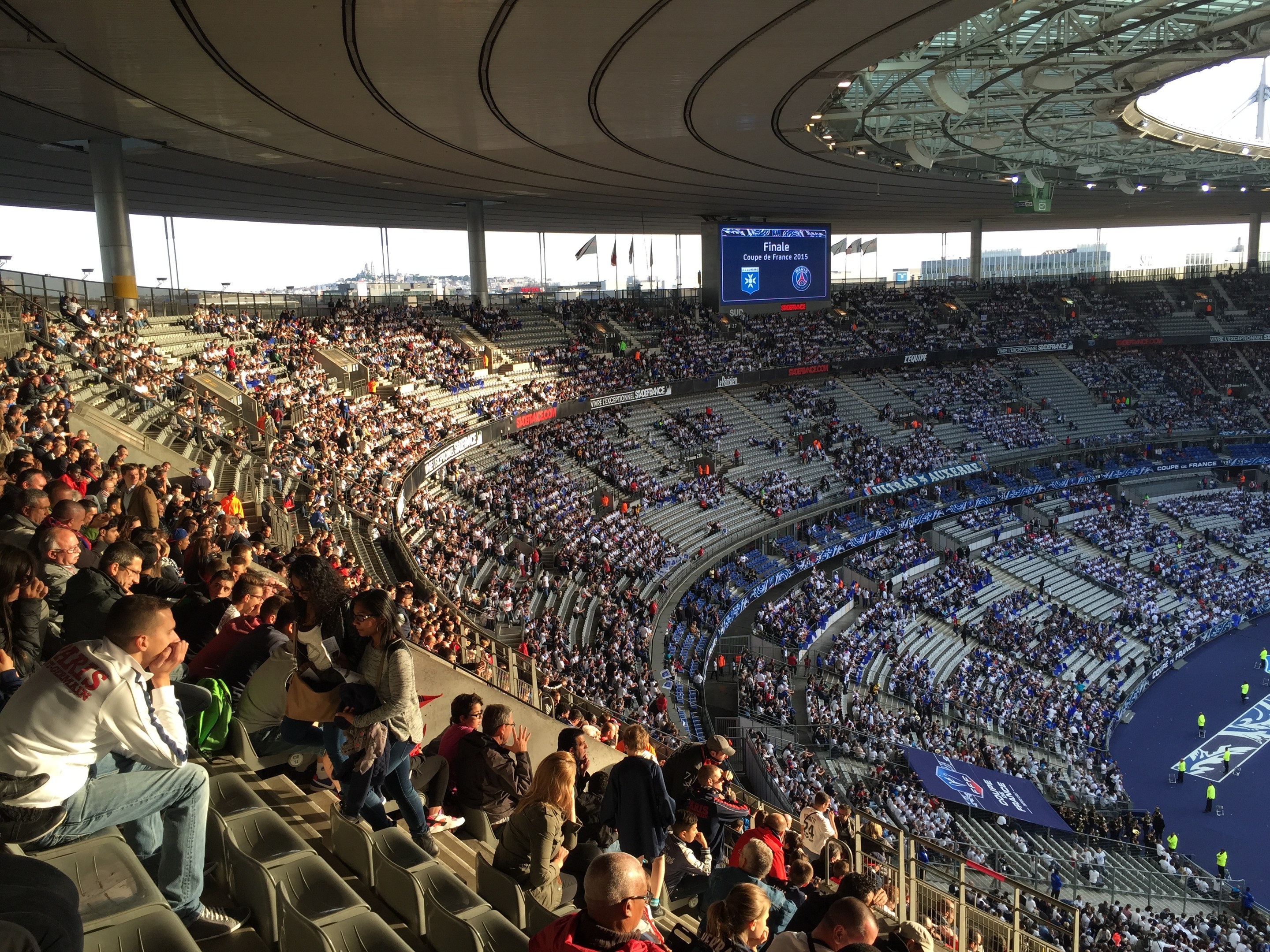 Große Menschenmenge in einem Stadion bei einem Förderballspiel mit einer Bühne, Fahnen, Masten und einem Bildschirm im Hintergrund, identifiziert als die Allianz Arena in München, Deutschland.
