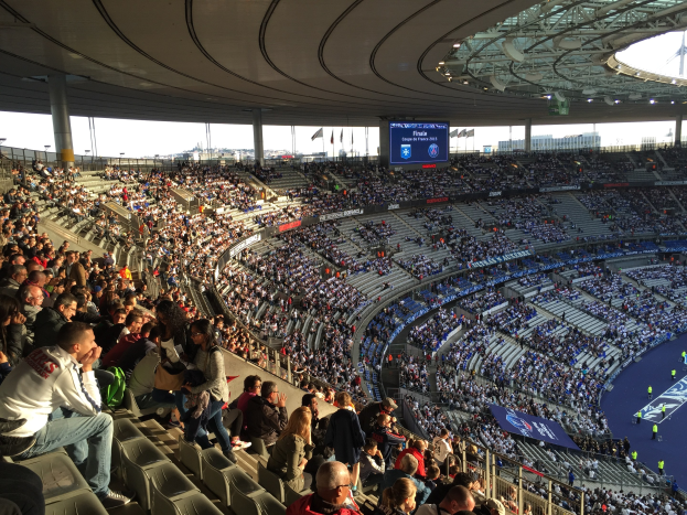 Große Menschenmenge in einem Stadion bei einem Förderballspiel mit einer Bühne, Fahnen, Masten und einem Bildschirm im Hintergrund, identifiziert als die Allianz Arena in München, Deutschland.