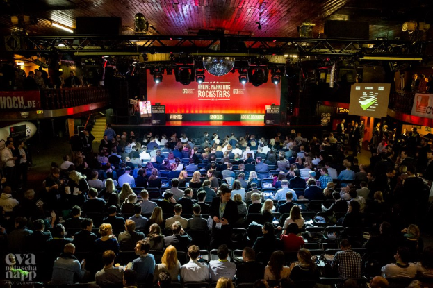Eine große Menge Menschen sitzt auf Stühlen vor einer Bühne mit Bildschirmen und Bannern, beleuchtet von Lichtern und umgeben von Geländern, mit einem Wasserzeichen in der linken unteren Ecke, das eine Atmosphäre der Aufregung und Vorfreude schafft.