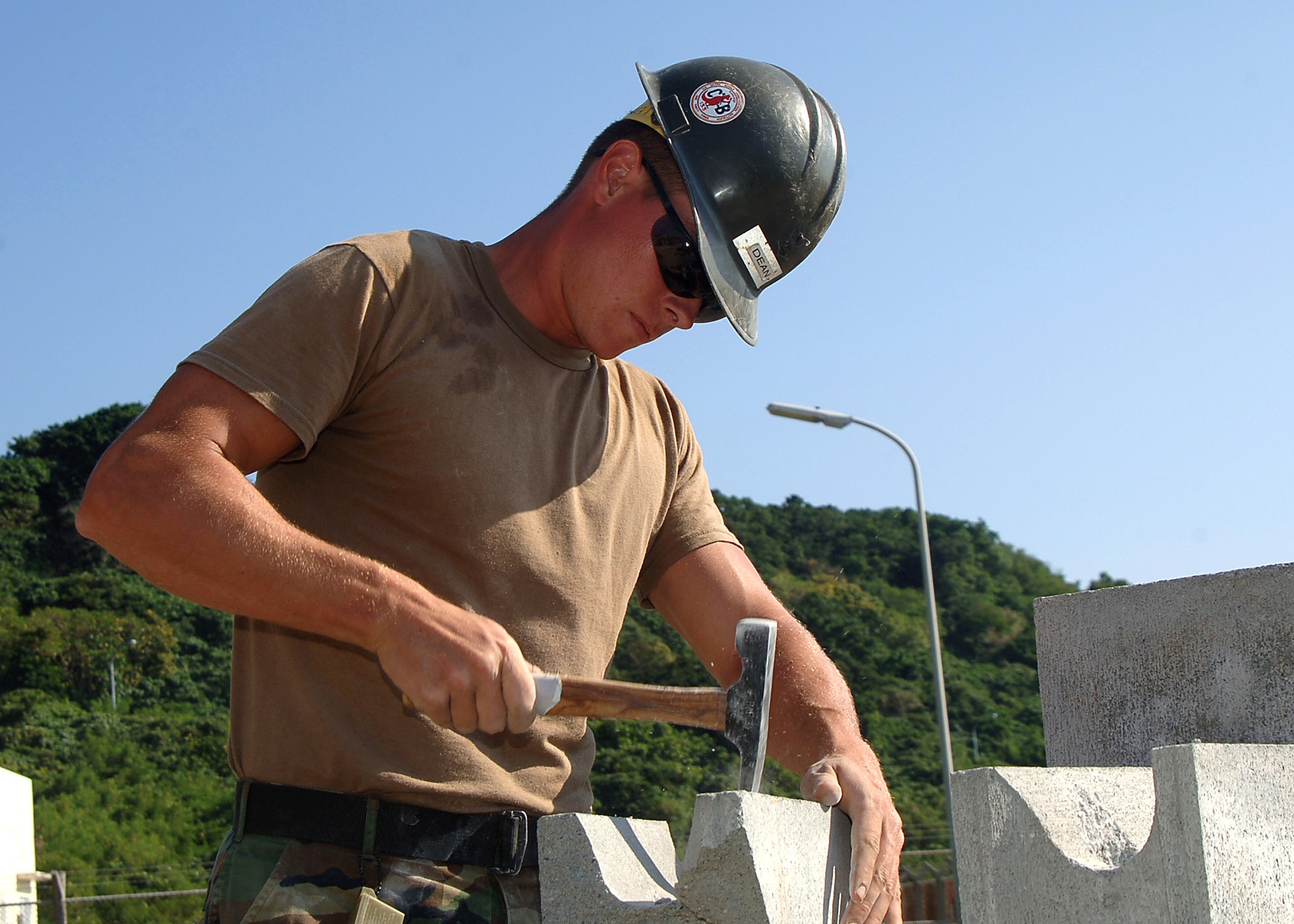 Ein Mann in einem Helm und einer Schutzbrille verwendet einen Hammer, um eine Wand im Freien zu bauen. Im Hintergrund sind Bäume, ein Laternenmast und ein klarer blauer Himmel zu sehen.
