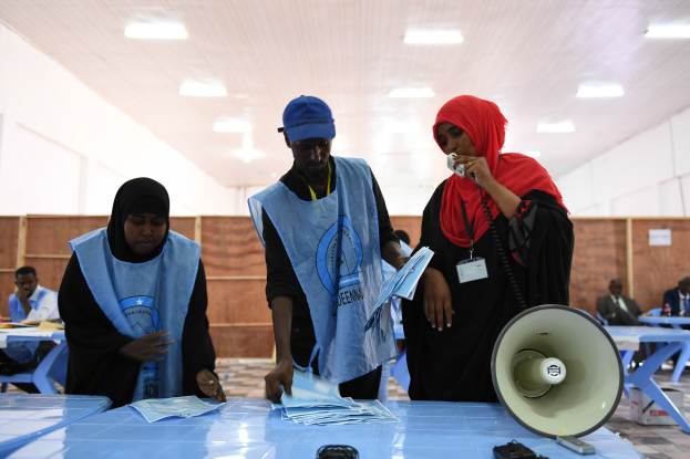Gruppe von Menschen um einen Tisch mit Megafon, Papieren und anderen Gegenständen an einer Wahllokal mit sitzenden und stehenden Personen im Hintergrund unter beleuchteter Decke.