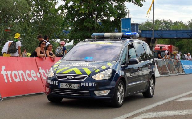 Polizeifahrzeug fahrt an einer Menge mit Schildern, Gel├Ąndern, B├Ąumen, einer ├Überquerung, einer Flagge und einem bew├Âlktem Himmel im Hintergrund vorbei.