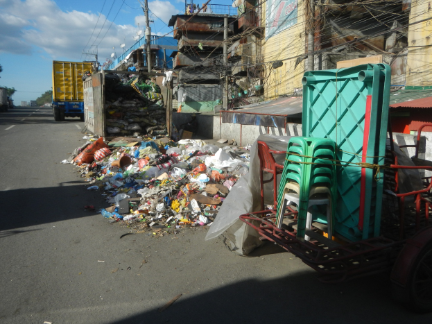 Ein Lastwagen neben einem Müllhaufen auf einer Straße, mit einem Wagen mit Plastikstühlen rechts daneben und Gebäuden, Strommasten, Bäumen und einem bewölkten Himmel im Hintergrund.