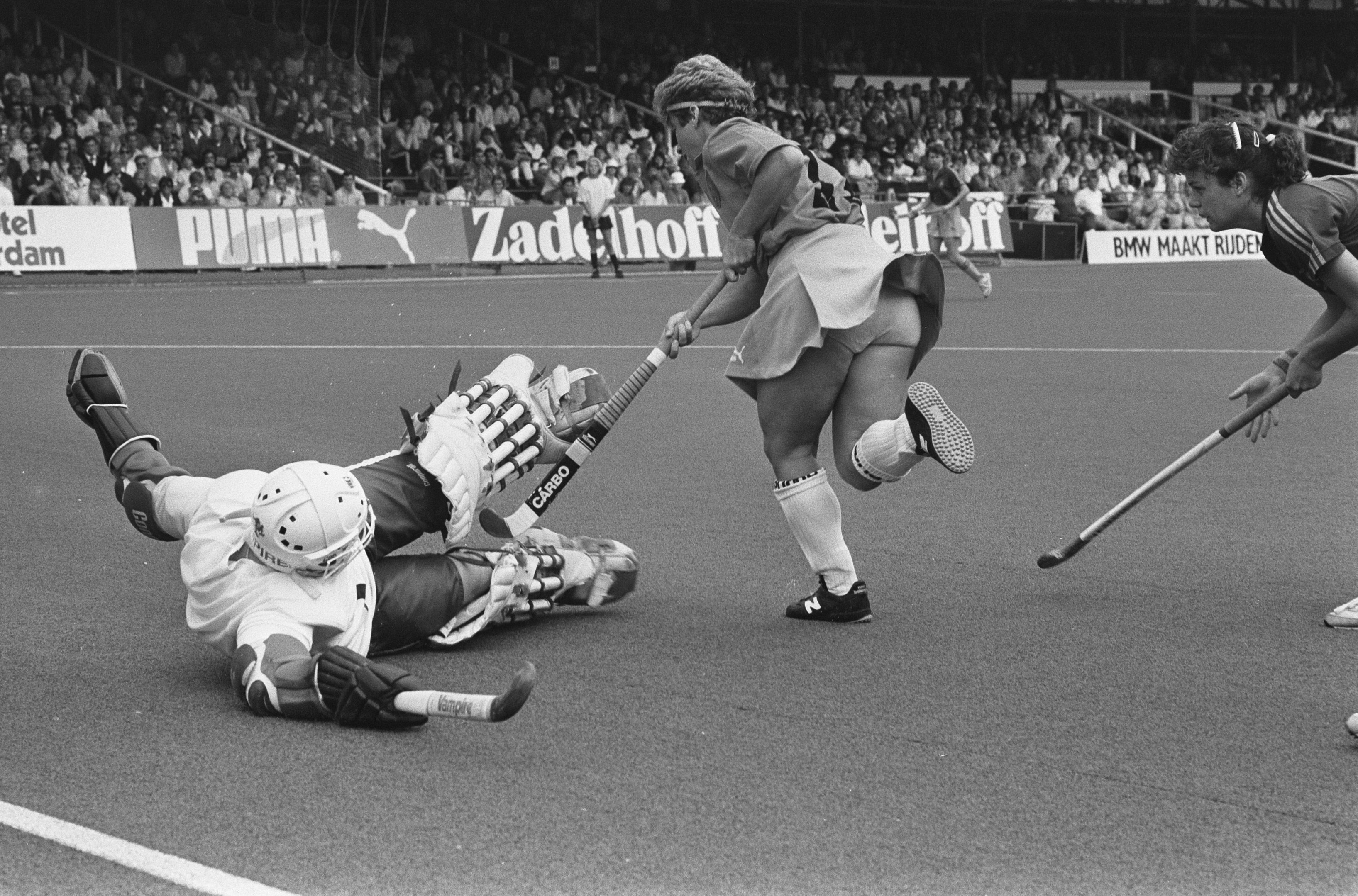 Schwarz-weißes Foto von Frauen, die Feldhockey in einem Stadion mit Zuschauern in den Rängen spielen.