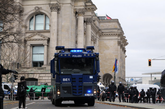 Gruppe von Polizisten vor einem großen Gebäude mit Fahrzeugen auf der Straße und einer Person mit einer Kamera links, mit Bäumen, Ampeln, Flaggen und einem klaren blauen Himmel im Hintergrund.