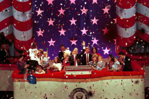 Eine Gruppe von Menschen auf einer Bühne mit einem Rednerpult, das ein Logo, Luftballons und Konfetti zeigt, vor einem Sternenhimmel und einem Vorhang, was auf eine Amtseinführung hindeutet.