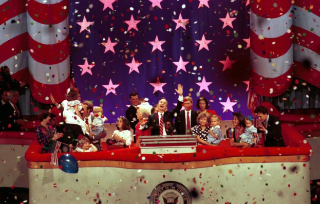 Eine Gruppe von Menschen auf einer Bühne mit einem Rednerpult, das ein Logo, Luftballons und Konfetti zeigt, vor einem Sternenhimmel und einem Vorhang, was auf eine Amtseinführung hindeutet.