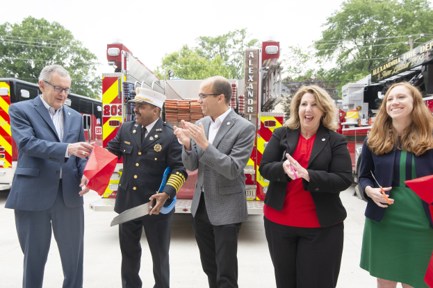 Group of people clapping and smiling at a ribbon cutting ceremony for the Alexandria Fire Department, with two individuals holding scissors and a red ribbon in front of a fire truck.