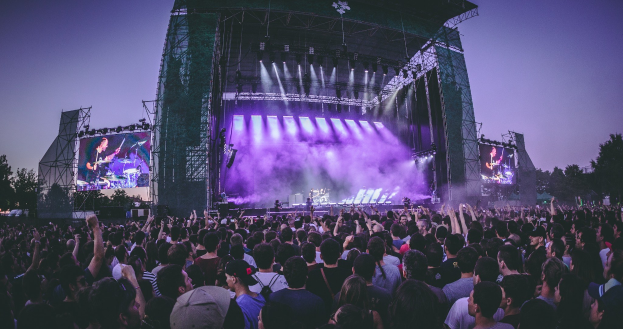 Eine große Menschenmenge bei einem Musikfestival schaut auf eine beleuchtete Bühne mit Instrumenten und Bildschirmen, die vor einem Gebäude, Bäumen und einem klaren blauen Himmel aufgebaut sind.