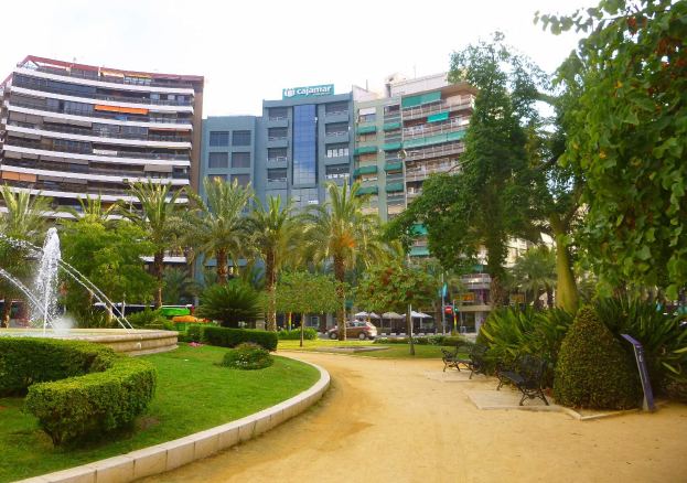 Ein Park mit einem zentralen Brunnen, umgeben von grünem Gras, Pflanzen, Bäumen, Bänken und Laternen, mit Fahrzeugen auf einer Straße und hohen Gebäuden im Hintergrund unter einem klaren blauen Himmel.