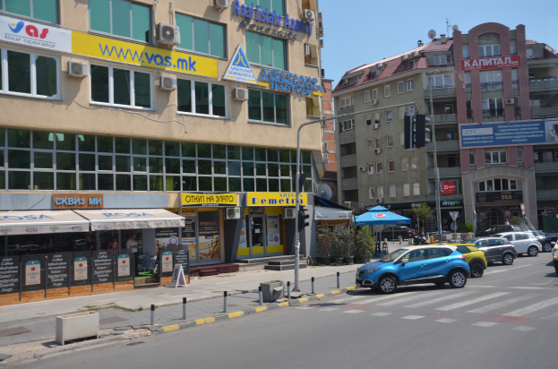 Eine Stadtstraße mit parkenden Autos, Gebäuden mit Fenstern, Schildern, einem Zelt, einer Gruppe von Menschen auf dem Gehweg, Topfpflanzen, einer Ampel und einem bewölkten Himmel.