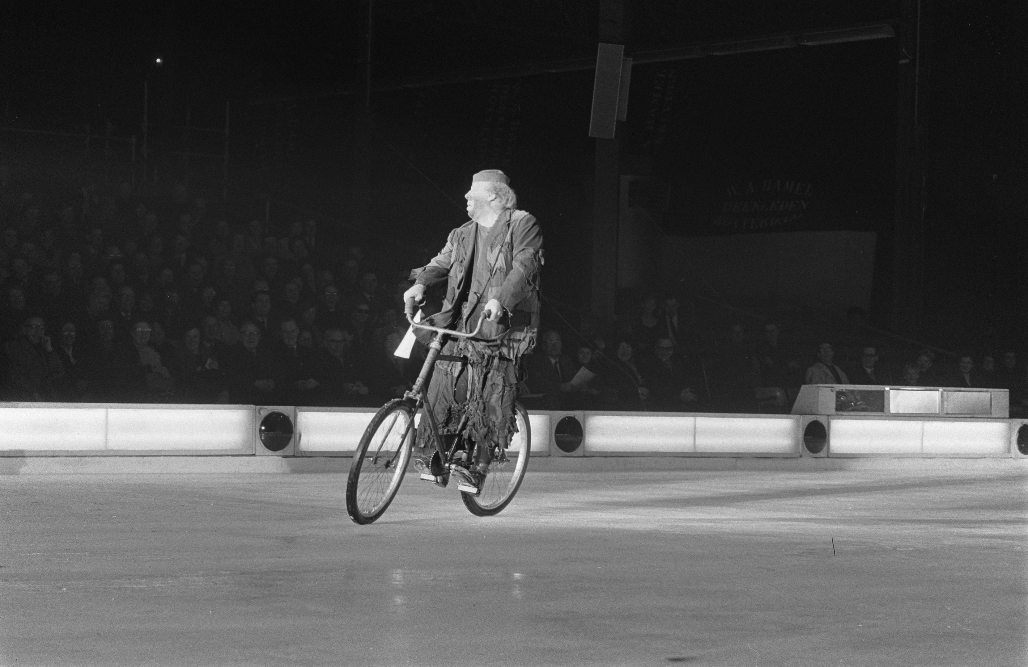 Schwarzes und weißes Bild eines Mannes, der auf einem Eisstadion Fahrrad fährt, mit einer Gruppe von Menschen im Hintergrund und einer Wand dahinter.