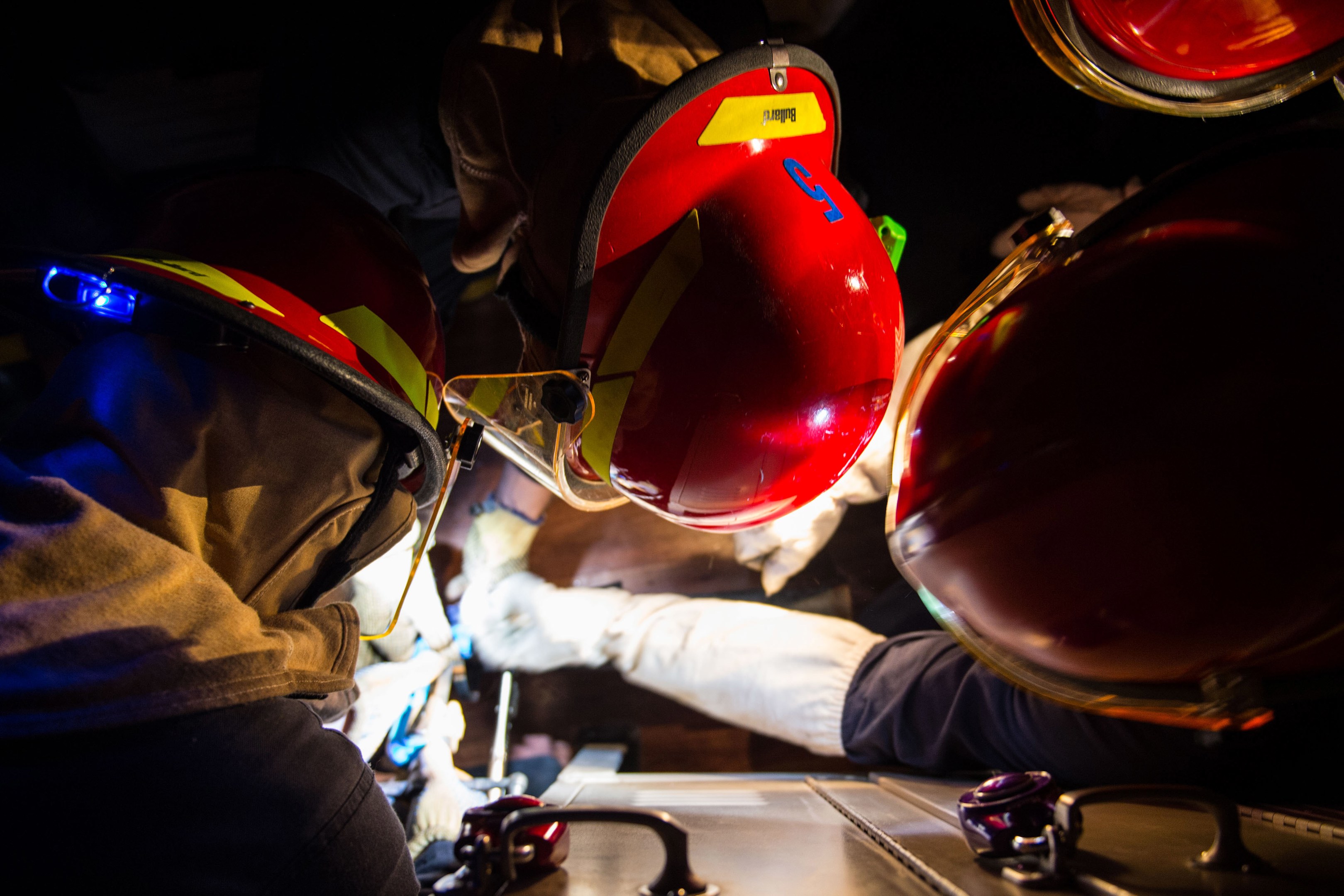 Feuerwehrleute in Helmen und Handschuhen arbeiten an einem Feuerwehrauto vor dunklem Hintergrund, mit einem Tisch mit Gegenständen auf der rechten Seite, die auf einen Notfall hinweisen.