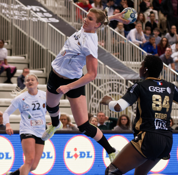 Eine Gruppe von Frauen, die auf einem Handballfeld spielen, mit einem Ball in der Luft, Zuschauern auf den Stufen und Geländern im Hintergrund und einem Schild mit der Aufschrift "Handball-Weltmeisterschaft 2019 - Tag 2".