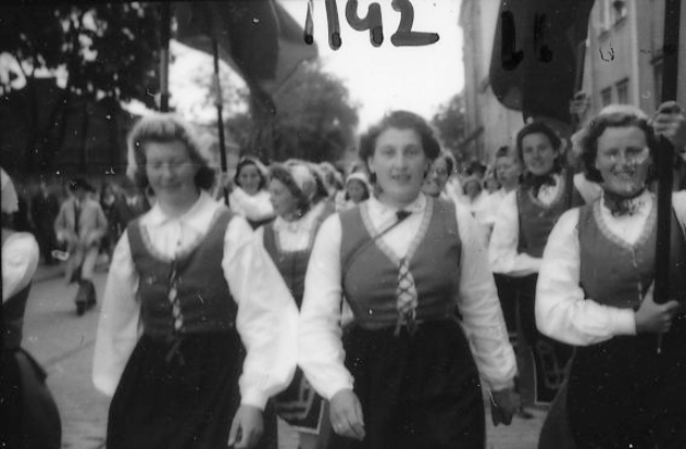 Eine Gruppe von Frauen in traditioneller bayrischer Tracht spaziert durch eine Straße, lachend, umgeben von Bäumen und Gebäuden, dargestellt in Schwarz-Weiß.