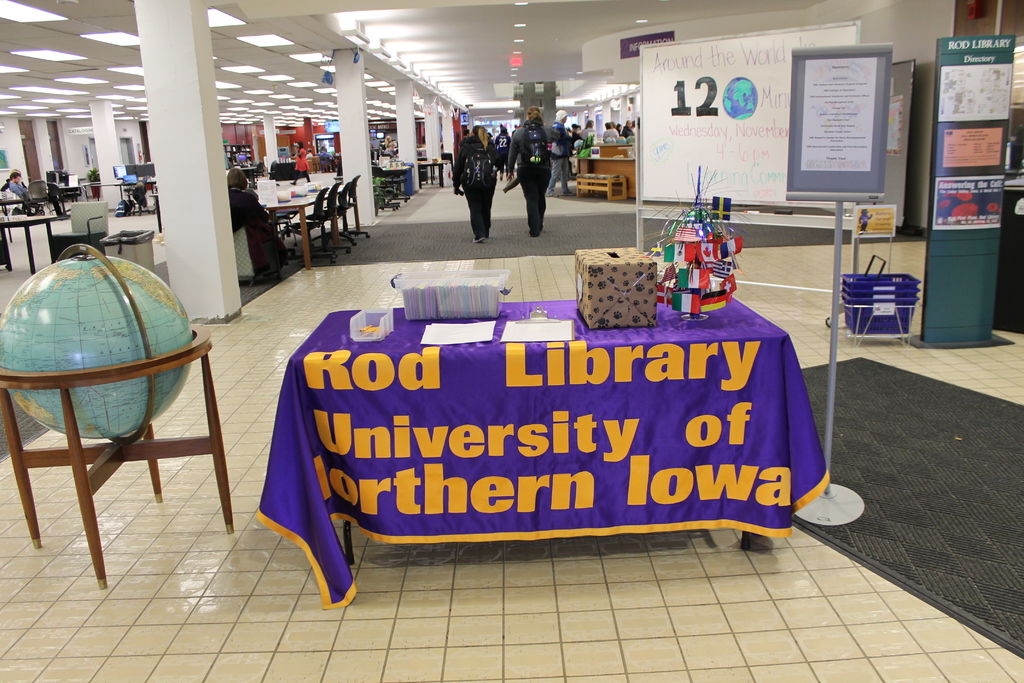 Eine Ausstellung mit einem Tisch, auf dem verschiedene Landesflaggen, eine Schachtel, Papiere und ein Banner sowie links daneben eine Weltkugel, rechts daneben eine Anschlagtafel mit einigen Menschen daneben und im Hintergrund viele Tische und Stühle zu sehen sind.