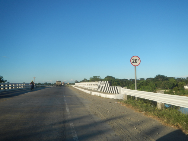 Fahrbahn mit Fahrzeugen, Tempolimit-Schild, Geländer, Gras, Bäume und klarer blauer Himmel.
