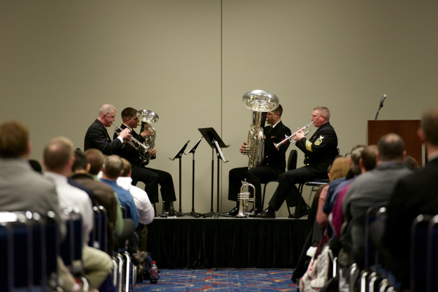 Eine Gruppe von Männern, die auf einer Bühne Blasinstrumente spielen, mit Notenständern, einem Rednerpult und einem Mikrofon vor einem sitzenden Publikum.