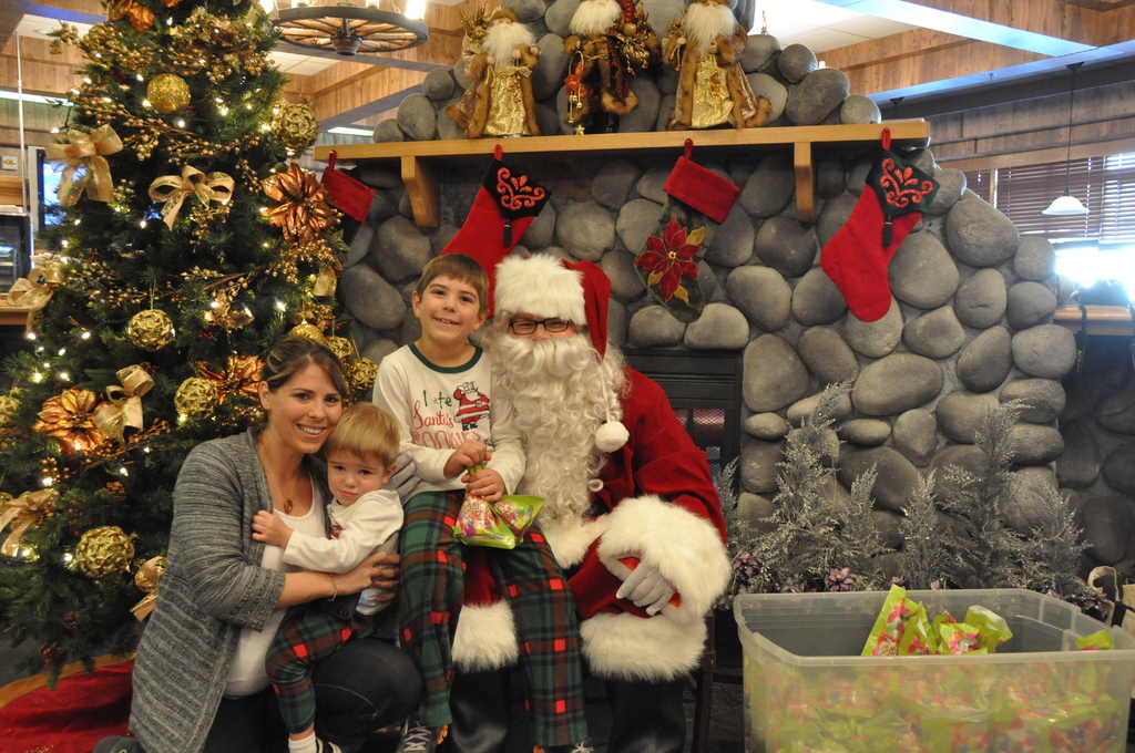 Eine Frau, Kinder und ein Mann in Santa-Claus-Kostüm mit Brille und Handschuhen stehen neben einem geschmückten Weihnachtsbaum, einer Wand mit Felsen, Spielzeugen, Dekorationen und einem Korb mit Gegenständen, mit Lichtern im Hintergrund.