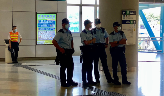 Eine Gruppe von Polizisten in Masken vor einem Gebäude mit Glastüren, Haftbefehle, eine Säule und ein Geländer, mit Bäumen und Fahrzeugen durch die Türen sichtbar.