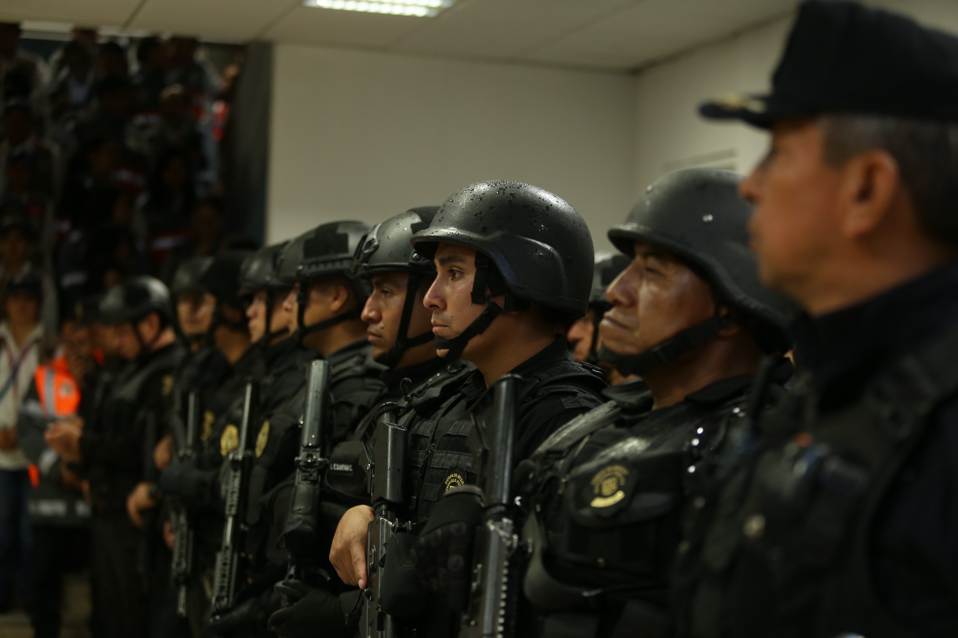Eine Gruppe von Polizisten in Helmen mit Waffen in der Hand, die neben einer Wand mit Deckenlampen stehen.