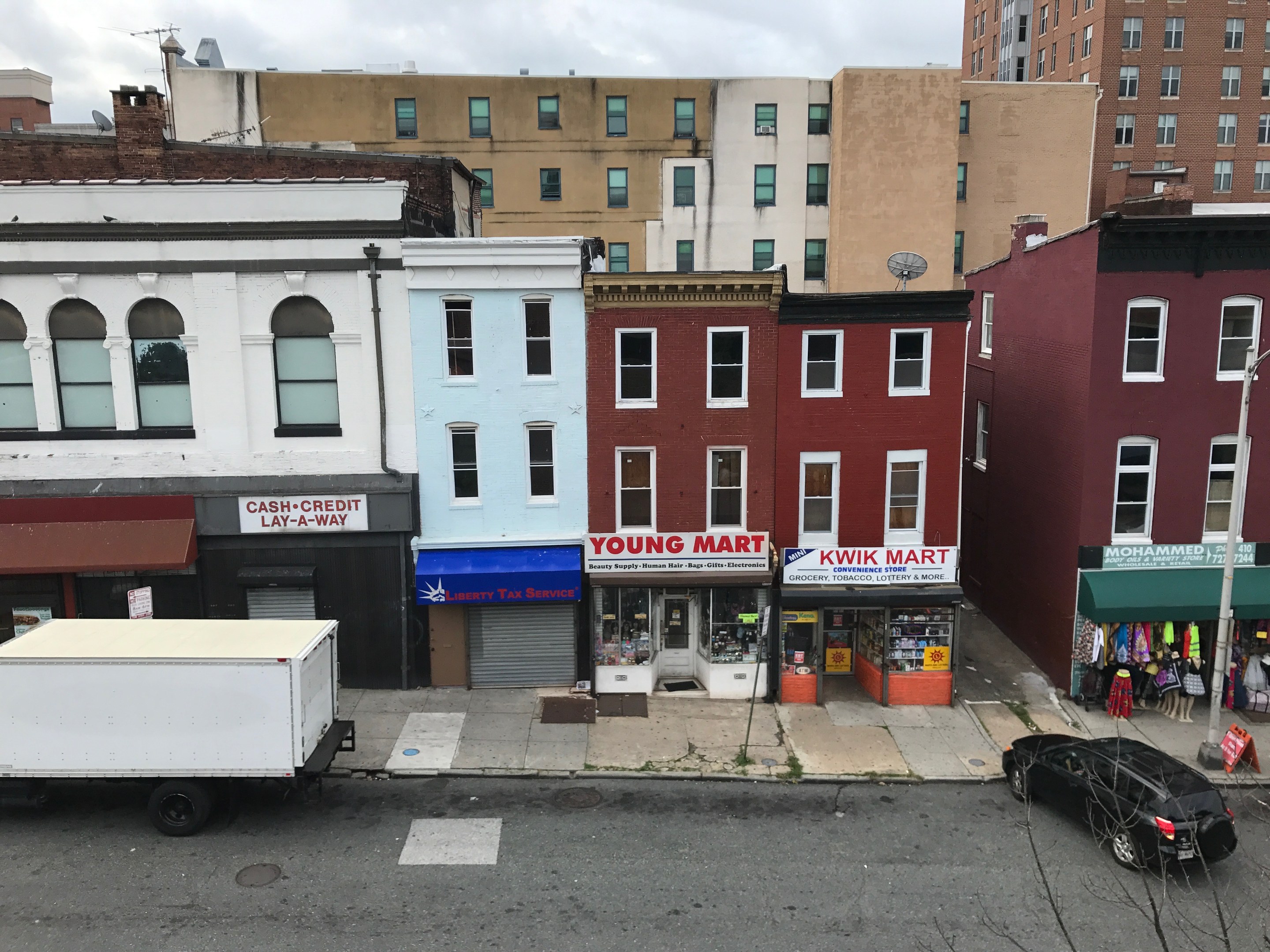 Luftaufnahme einer belebten Straße in Brooklyn, New York, mit Fahrzeugen, Geschäftsfronten, Werbetafeln, Strommasten und Drähte vor einem bewölkten Himmel, was auf ein kommerzielles Grundstück zum Verkauf hinweist.