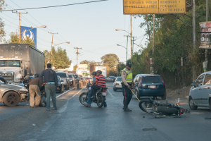 Eine Gruppe von Menschen um ein verunglücktes Motorrad auf der Straße herumstehend mit mehreren Fahrzeugen, darunter ein Lastwagen, und einem Hintergrund aus Bäumen, Polen, Lampen und Schildern unter dem Himmel.