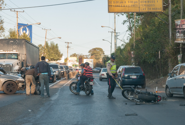 Eine Gruppe von Menschen um ein verunglücktes Motorrad auf der Straße herumstehend mit mehreren Fahrzeugen, darunter ein Lastwagen, und einem Hintergrund aus Bäumen, Polen, Lampen und Schildern unter dem Himmel.
