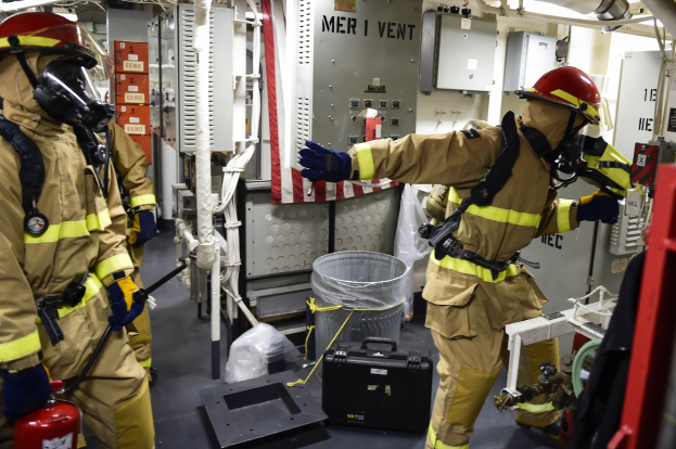 Drei Feuerwehrleute in Schutzausrüstung stehen neben einem Feuerlöscher, mit einer Maschine, Rohren und verstreuten Gegenständen im Hintergrund.