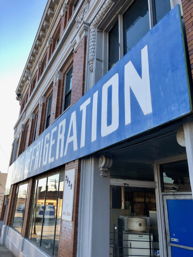 Ein Gebäude mit Glaswänden und einer blauen Tafel, einem sichtbaren Kühlschrank im Inneren und einem Himmelhintergrund, wahrscheinlich ein Kühlgeräteladen.