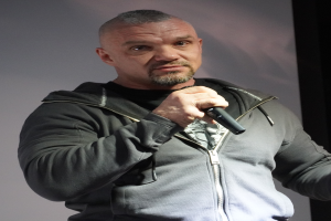 Ein Mann in einer schwarzen Kapuzenjacke, schwarzem Anzug und weißem Hemd mit zurückgek├Ąmmtem Haar h├Ąlt ein Mikrofon und steht selbstbewusst vor einer Wand.